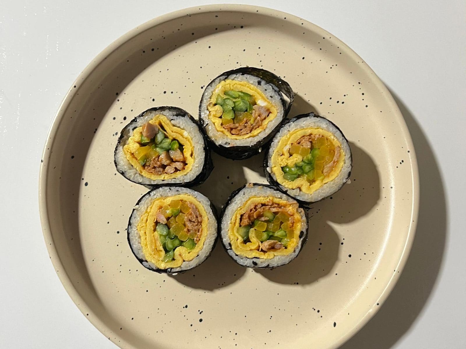 Five thick slices of Korean kimbap on a cream plate with black speckles, each slice showing white rice, a dark seaweed wrap, a yellow omelette layer, green cucumber, yellow pickled radish and seasoned meat.