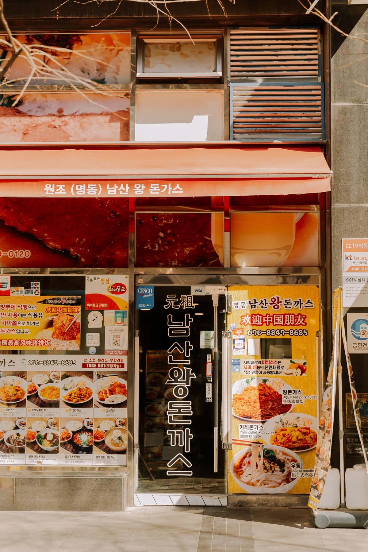 The front of a tonkatsu restaurant in Myeongdong, Seoul, with an orange awning, a vertical Korean sign and windows covered in picture menus, including a yellow board that welcomes Chinese visitors in Chinese.