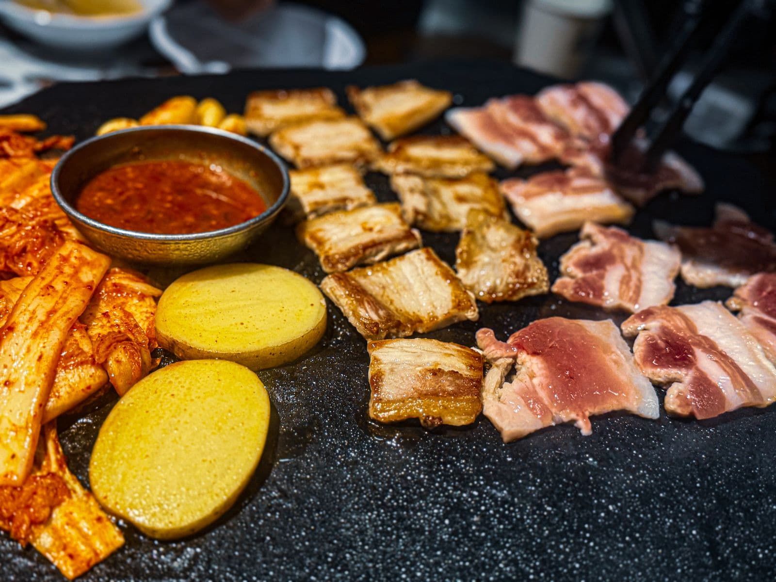 Grilled pork belly slices beside raw ones on a black stone grill plate, with kimchi, round potato slices and a small metal bowl of red dipping sauce.